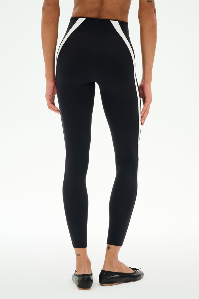 Model wears SPLITS59 Miles High Waist Rigor 7/8 leggings in Black/White, featuring white side panels and black flat shoes, standing with back to camera against a plain white background.