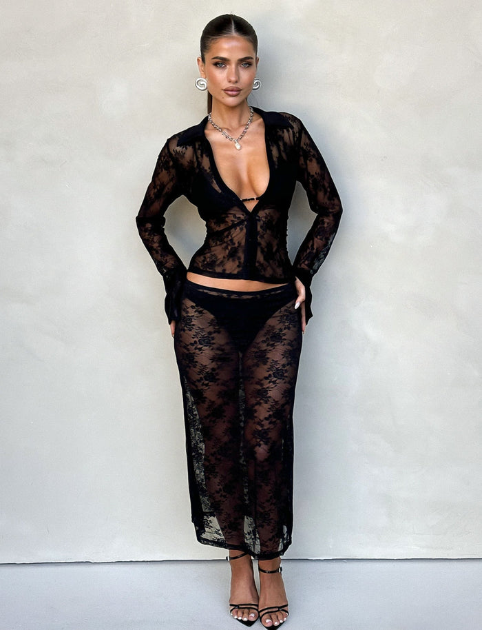 A woman stands against a beige wall wearing TIGERMIST's MIRABEL SKIRT in black lace, featuring a sheer long-sleeved top and skirt with visible black underwear beneath. She accessorizes with hoop earrings, a necklace, and black high-heeled sandals, with her hair styled in a sleek, pulled-back look.