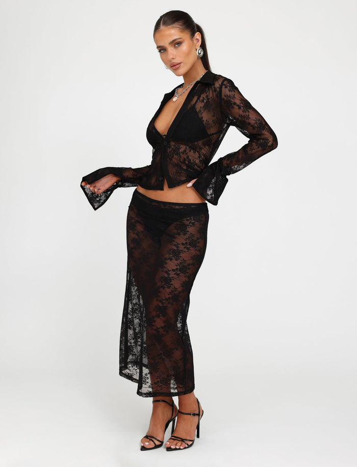 A woman is posing against a plain white background, wearing the sheer black MIRABEL SKIRT by TIGERMIST with a matching bra and underwear underneath. She is accessorized with statement earrings, a necklace, and black high-heeled sandals. Her hair is styled back.