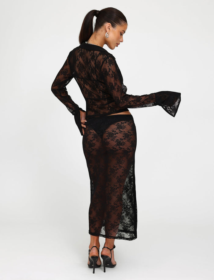 A woman stands in a studio, facing away from the camera, wearing a TIGERMIST black lace long-sleeve top and the matching MIRABEL SKIRT - BLACK. Her hair is tied in a ponytail and she is wearing black high-heeled sandals.