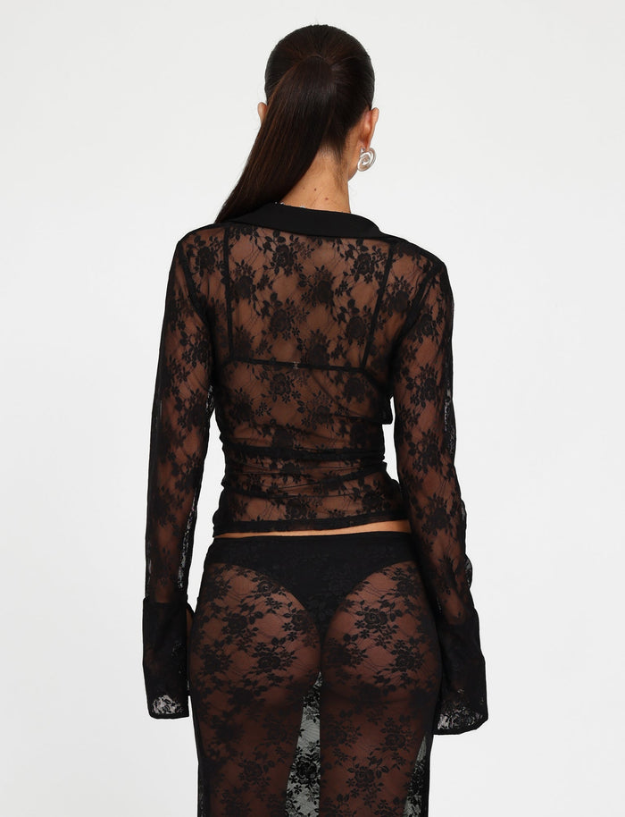 Seen from the back, a person with long hair tied in a ponytail is wearing the MIRABEL TOP - BLACK and matching lace pants by TIGERMIST. Both garments are sheer black lace, revealing a black undergarment underneath. The background is plain white.