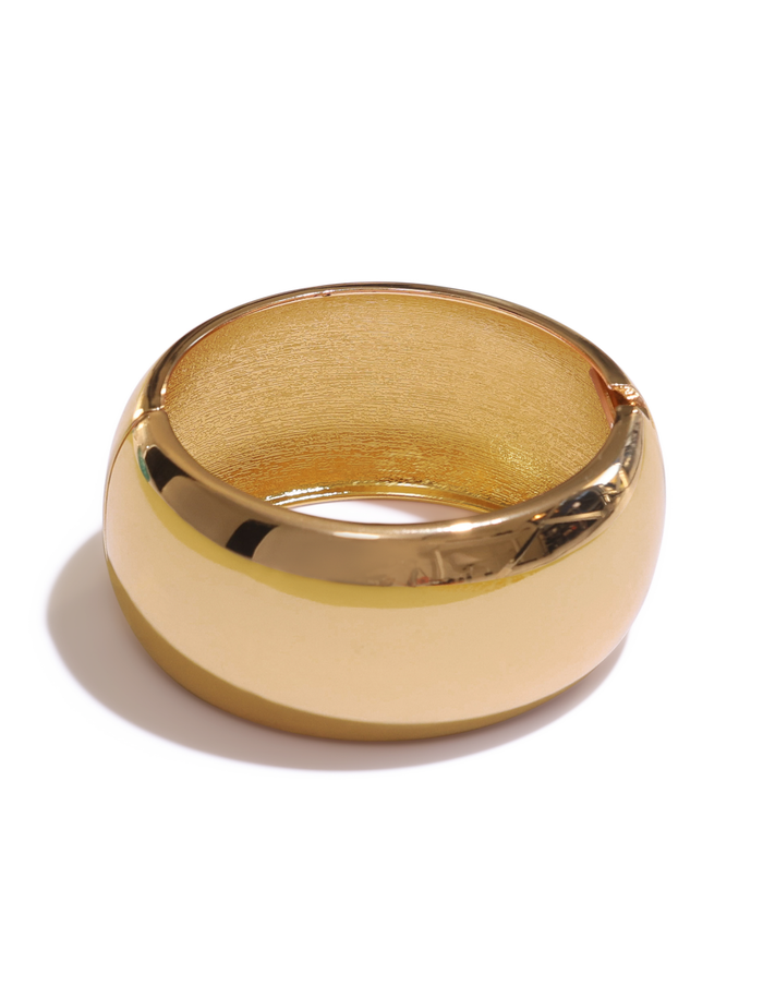 The TIGERMIST MIRAH CHUNKY BANGLE - GOLD is a wide, polished gold bangle with a smooth, shiny surface, reminiscent of a gold cuff bracelet and casting a soft shadow on a white background.