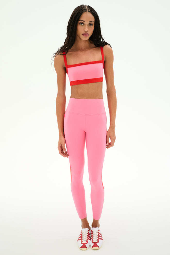Wearing a SPLITS59 Monah Rigor Bra in Strawberry/Lotus Red, matching high-waisted leggings, and red-and-white sneakers, the person stands against a plain white background.