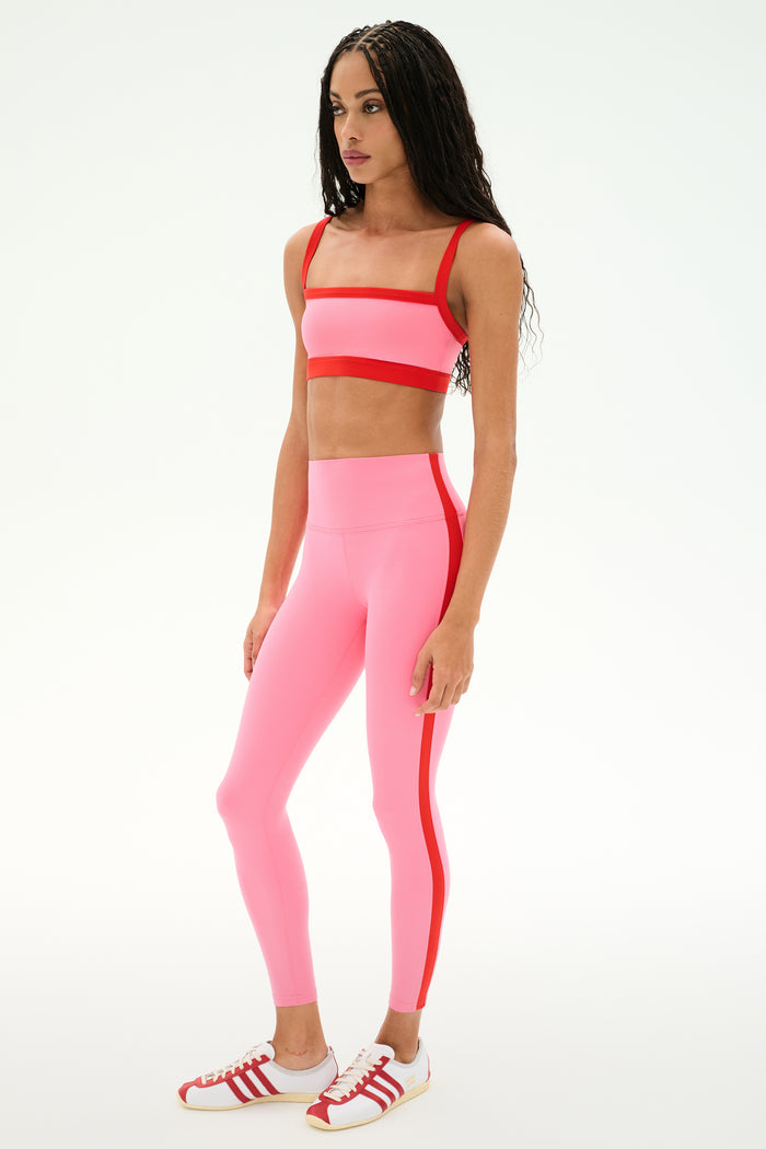 A woman stands in a studio wearing the SPLITS59 Monah Rigor Bra in Strawberry/Lotus Red, paired with high-waisted pink leggings featuring a red stripe and white sneakers with red accents.