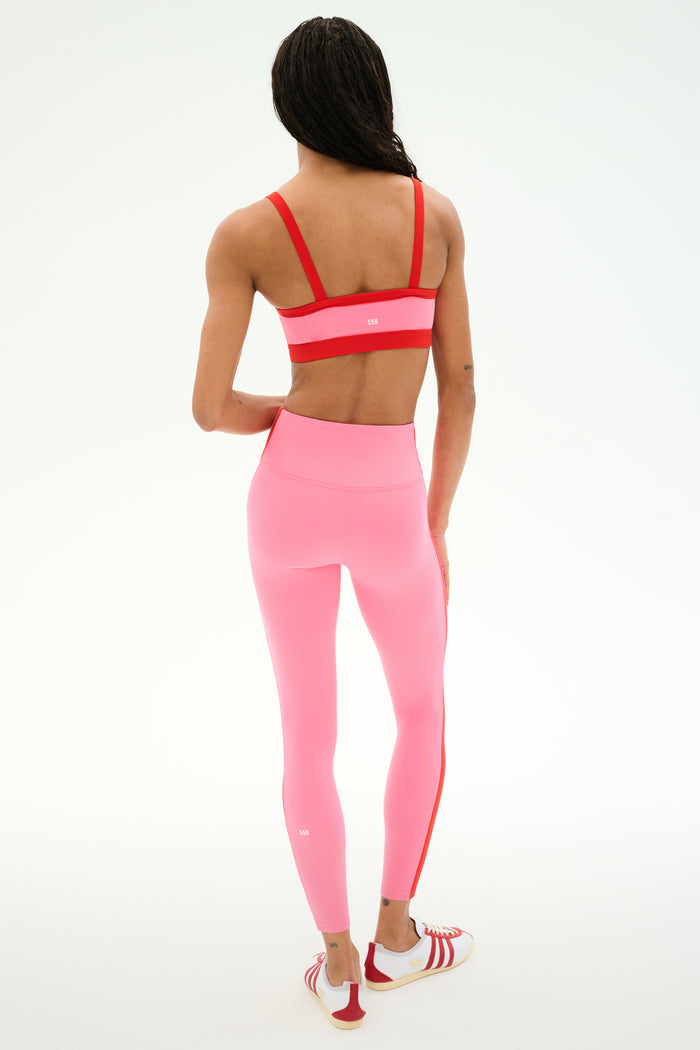 A person stands facing away, wearing a SPLITS59 Monah Rigor Bra in Strawberry/Lotus Red, high-waisted pink leggings, and red-and-white athletic shoes against a plain white background.