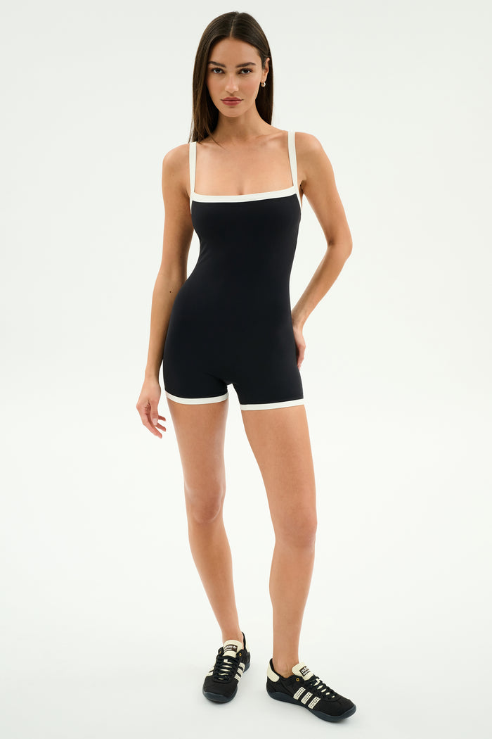 A woman stands against a plain background wearing the SPLITS59 Monah Rigor Jumpsuit in black/white—made from supportive Rigor fabric for high-impact workouts—and black sneakers with white stripes.