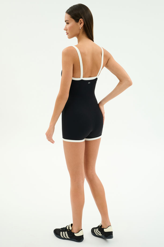 A woman faces away, wearing the SPLITS59 Monah Rigor Jumpsuit in Black/White with black Adidas sneakers, against a plain white background.