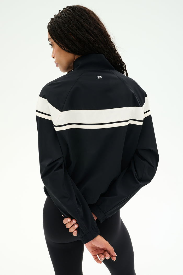 A person stands with their back to the camera, wearing the SPLITS59 Morrison Nylon Twill Track Jacket in Black/Creme, featuring a white stripe across the back, paired with black spandex leggings. Their hands are clasped behind their back.