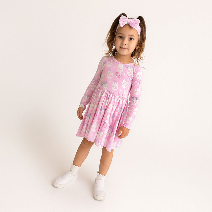 Child standing, wearing the Posh Peanut Ms. Kelly Bow Back Twirl Dress, pink with white skull print, white background