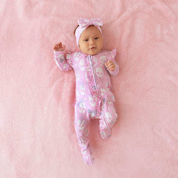 Baby lying down, wearing the Posh Peanut Ms. Kelly Luxe Bow Headwrap, pink background
