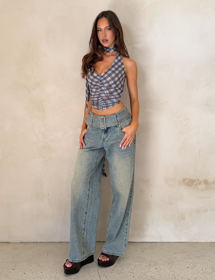 A woman poses against a light textured wall, wearing the TIGERMIST NOBLE TOP in blue check, paired with light-wash wide-leg jeans with a belt, dark platform sandals, and a matching neck scarf.