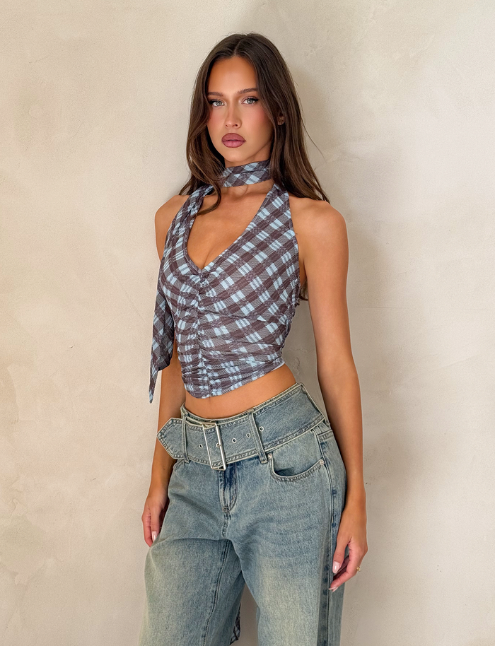 A woman with long brown hair wears the TIGERMIST NOBLE TOP - BLUE CHECK with loose high-waisted jeans and a wide belt, standing against a beige textured wall.