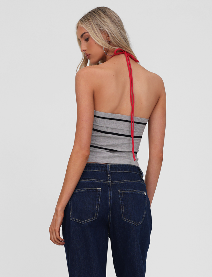 A woman with long blonde hair is seen from behind, wearing the TIGERMIST NOUR TOP in grey stripe and dark blue jeans, standing against a plain light background.
