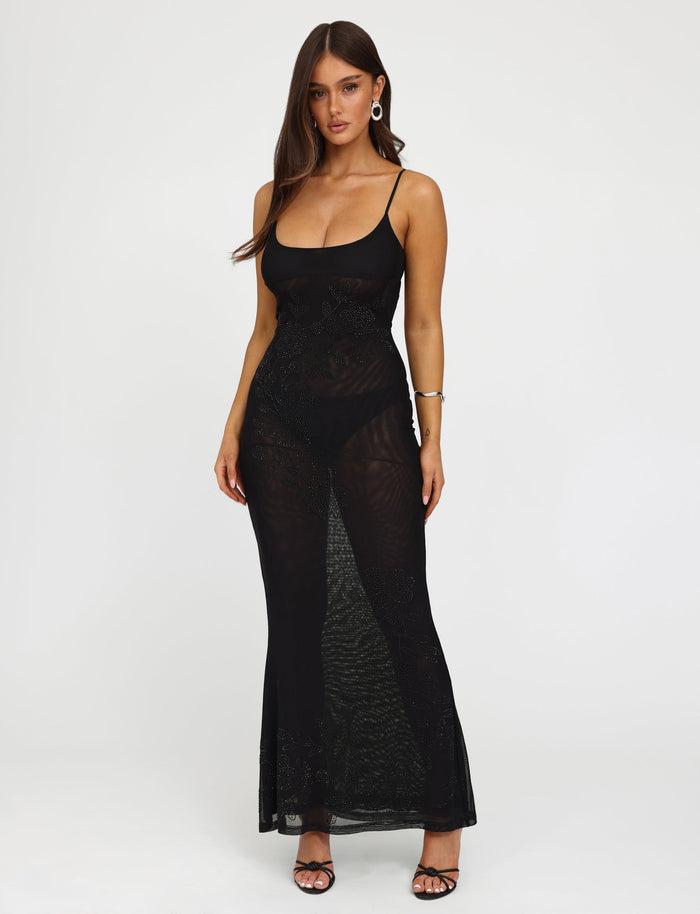 A woman with long hair wears the OCTAVIA DRESS - BLACK from TIGERMIST, a sleeveless, form-fitting, sheer black dress featuring intricate patterns throughout. She pairs the dress with black high-heeled sandals, hoop earrings, and a bracelet on her left wrist. The background is plain white.
