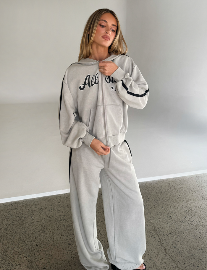 A woman stands indoors on a concrete floor wearing the TIGERMIST PAYTON HOODIE in stone with a matching tracksuit, looking slightly to the side and touching her collar.