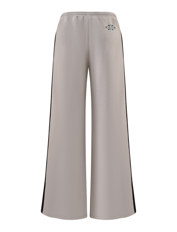 The PAYTON TRACKPANT - STONE by TIGERMIST features a wide-leg fit, taupe cotton fabric, elastic waistband, black side panels, and small black symbols near the back waist. The sporty pants are shown against a white background.