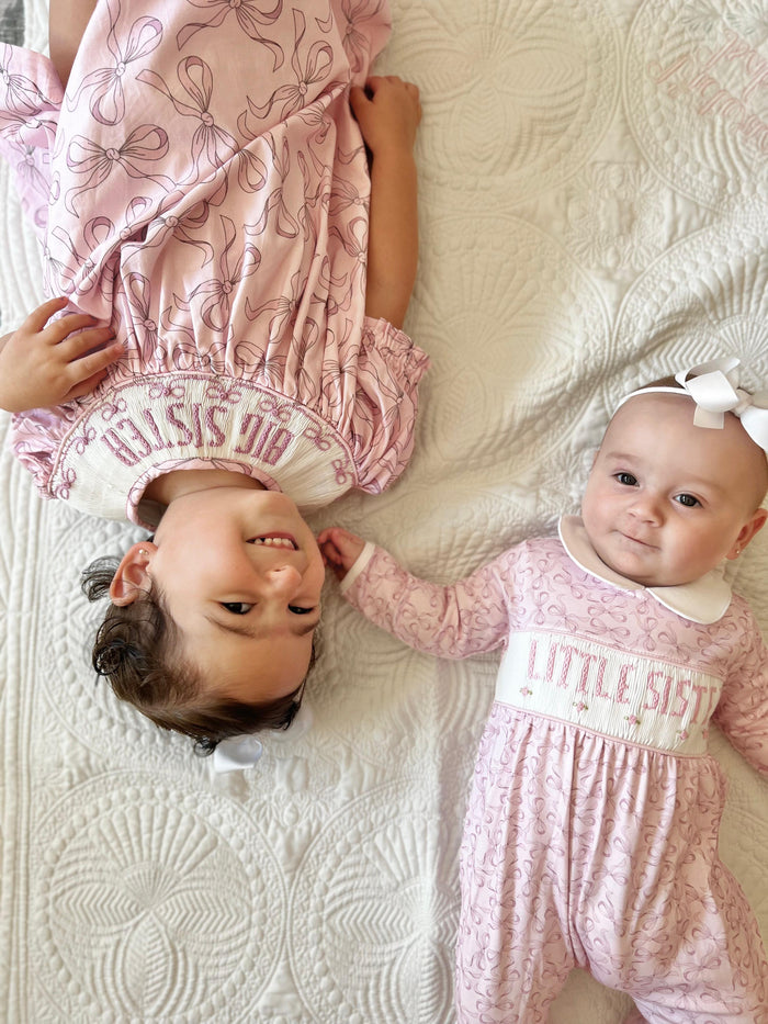 Two girls wearing matching Little Sister Bows Footie Babygrows in soft pink bow print with handsmocked lettering