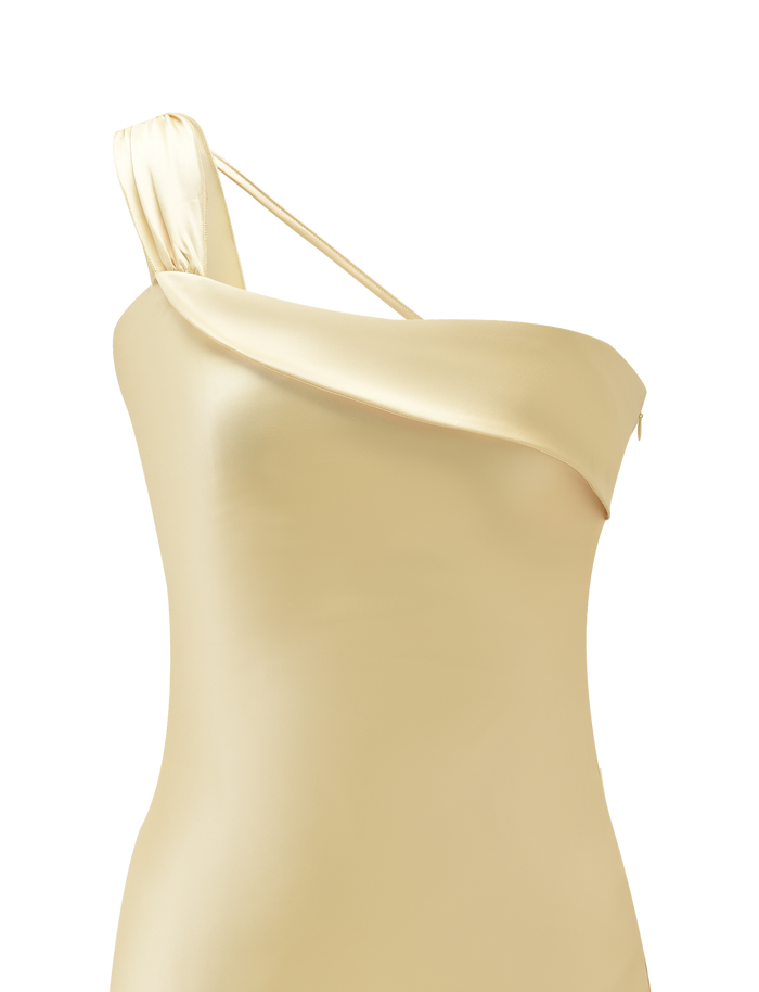 A close-up of the TIGERMIST RAINEY MINI DRESS in Butter, featuring a light yellow satin fabric, one-shoulder design, asymmetrical neckline, and draped detail. The background is plain white.