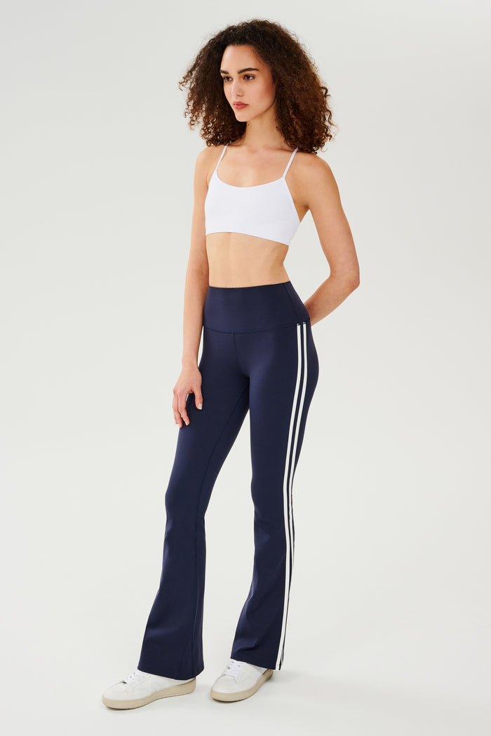Full front view of woman with dark curly hair wearing dark blue high waist below ankle length legging with wide flared bottoms and white double side stripes and white bra with spaghetti straps. Paired with white shoes.