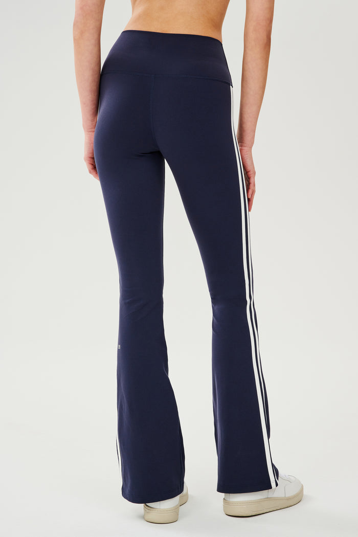 Back view of woman wearing dark blue high waist below ankle length legging with wide flared bottoms and white double side stripes on both legs and white S59 logo on back of left calf and white and side black stripes on both legs. Paired with white shoes.