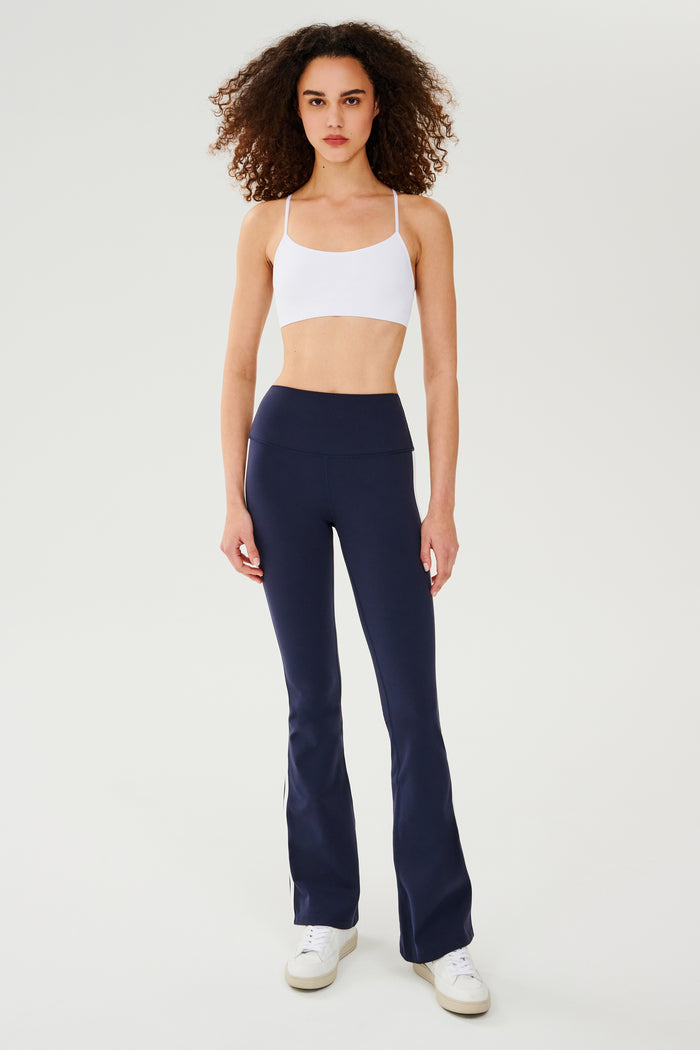 Full front view of woman with dark curly hair wearing dark blue high waist below ankle length legging with wide flared bottoms and white double side stripes and white bra with spaghetti straps. Paired with white shoes.