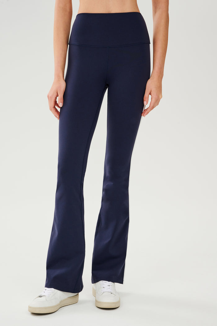 Front view of woman wearing dark blue high waist below ankle length legging with wide flared bottoms and white double side stripes on both legs and white S59 logo on back of left calf and white and side black stripes on both legs. Paired with white shoes.