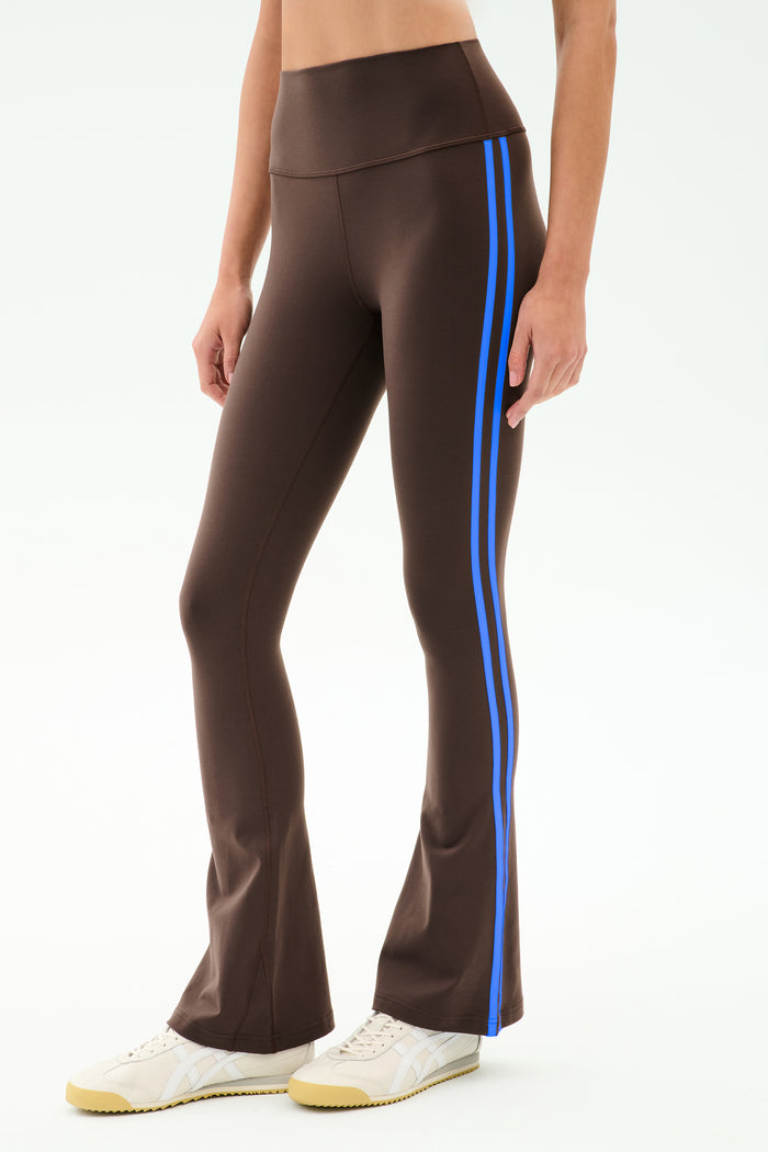 Model wears the SPLITS59 Raquel High Waist Supplex Flare in Dark Chocolate/Classic Blue, featuring blue side stripes and a comfy 4-way stretch fabric—ideal for gym workouts—paired with white sneakers against a plain backdrop.