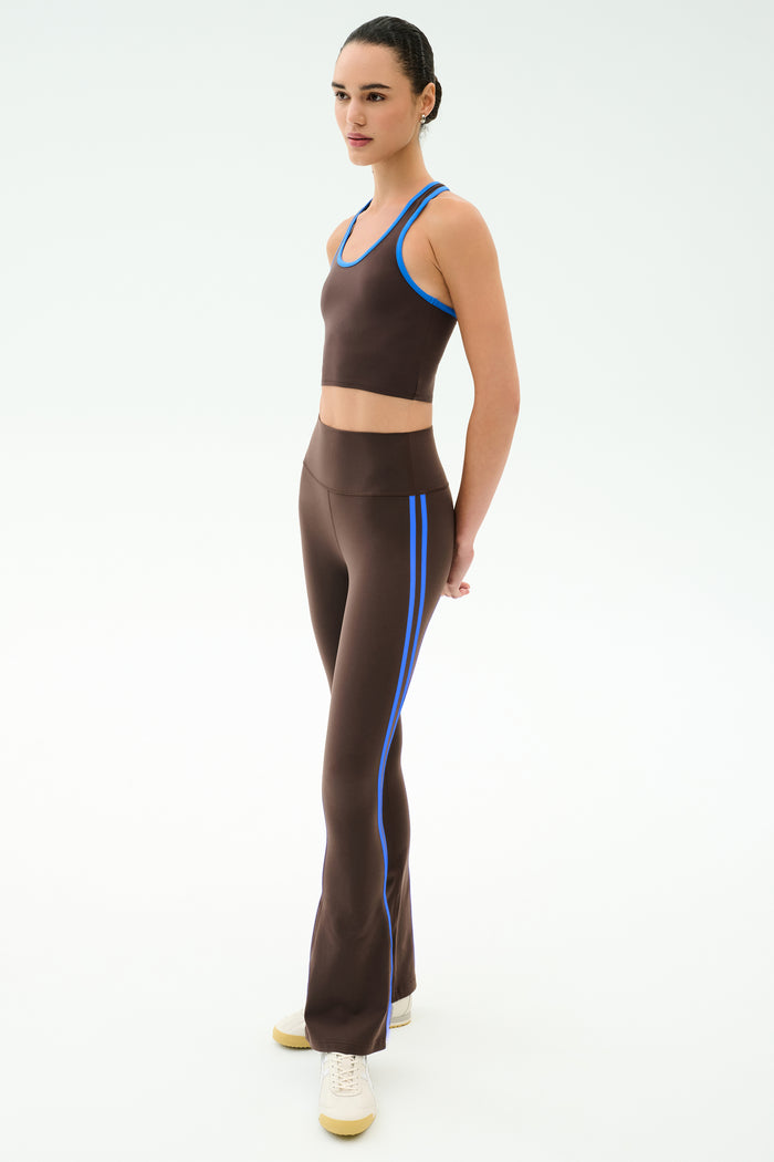 A woman wears the SPLITS59 Raquel High Waist Supplex Flare in Dark Chocolate/Classic Blue with matching crop top and cream sneakers, against a plain light background.
