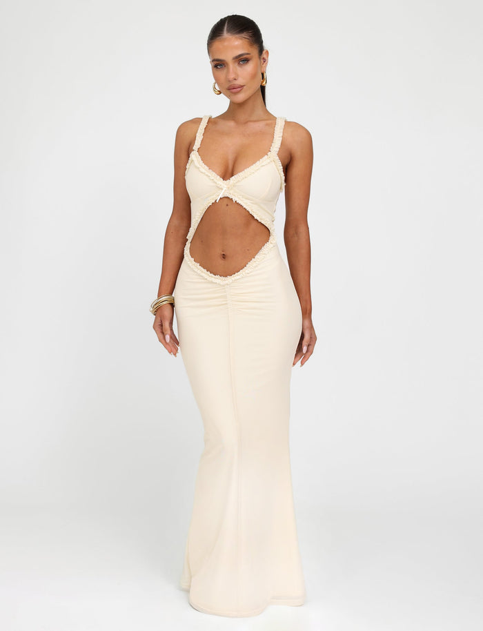 Wearing the RAYLA DRESS - WHITE : CREAM from TIGERMIST, a woman showcases its long, form-fitting silhouette with a unique midsection cutout design, spaghetti straps, and plunging neckline. Her long hair is pulled back as she accessorizes with hoop earrings and bangles, standing against a plain background.