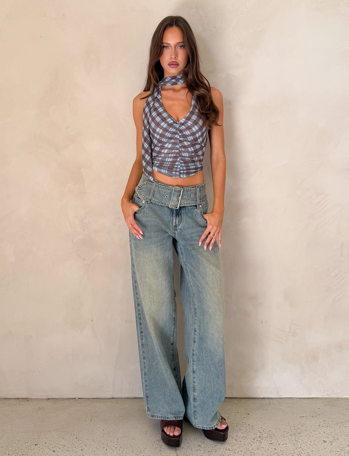 A woman stands against a light beige wall, wearing a blue checkered criss-cross halter top, black platform sandals, and TIGERMIST REEF JEAN - LIGHTWASH vintage wash boyfriend jeans with a belt in a relaxed fit.