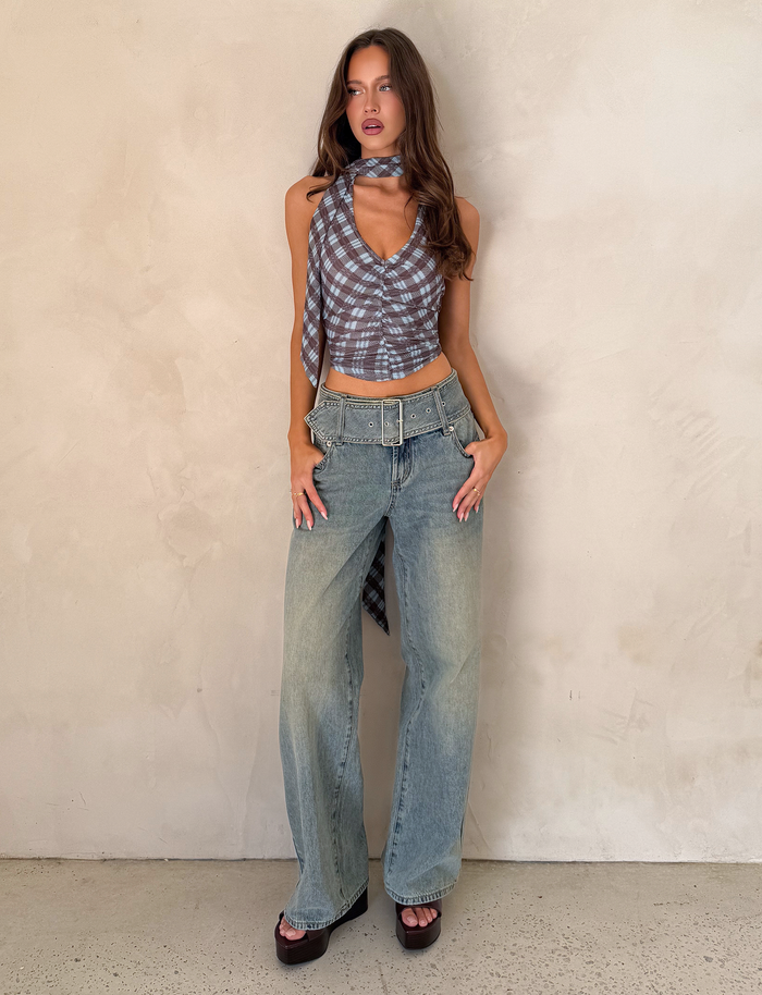 A woman with long wavy hair strikes a confident pose against a light wall, wearing TIGERMIST REEF JEAN - LIGHTWASH, a sleeveless plaid crop top with scarf detail, wide belt, and dark platform sandals.