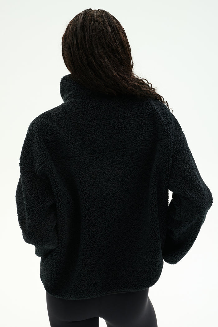 A person stands with their back to the camera, wearing the SPLITS59 Reese Sherpa Half Zip Pullover in black—crafted from 100% polyester—paired with black leggings, set against a plain white background.