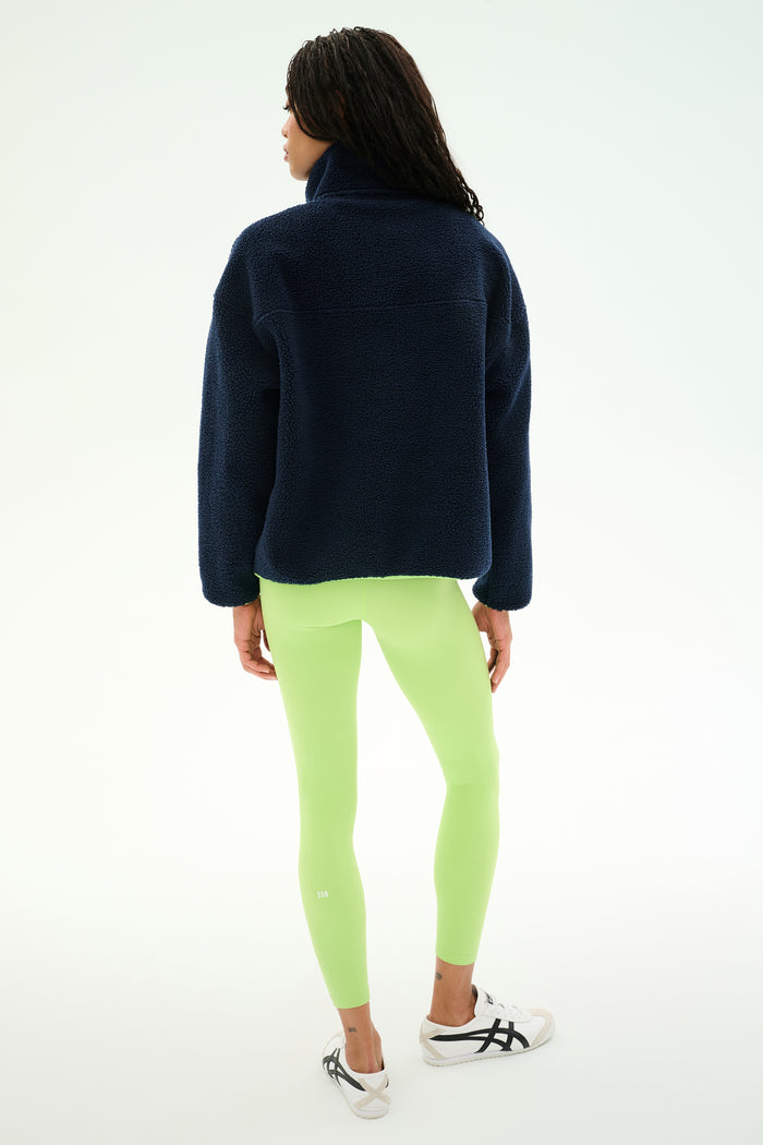 A woman stands with her back to the camera, wearing the SPLITS59 Reese Sherpa Half Zip Pullover in Indigo/Limeade, paired with neon green leggings and white sneakers against a plain light background.