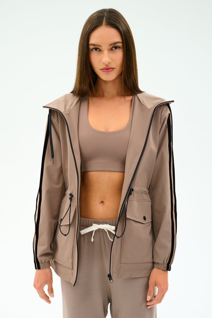 A woman with straight brown hair wears the SPLITS59 Reggie Water Repellent Jacket in Latte/Black, styled with matching taupe sportswear, against a plain white background.