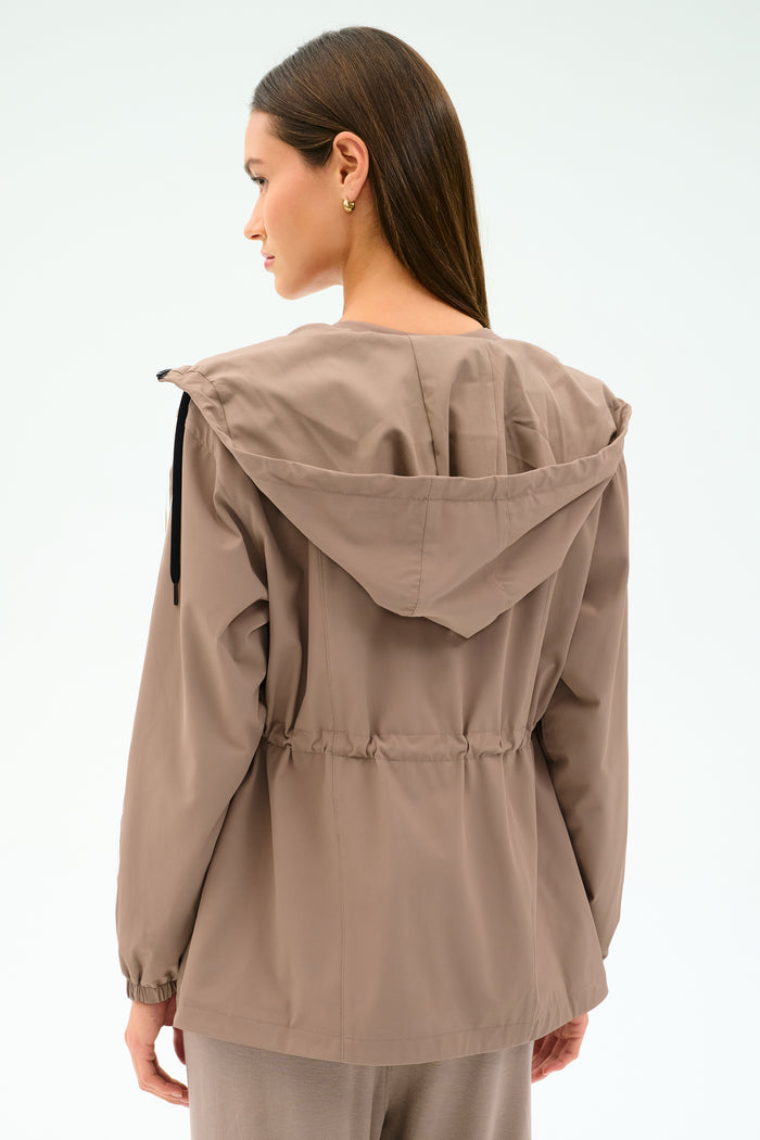 A woman with long brown hair is seen from the back wearing the SPLITS59 Reggie Water Repellent Jacket in Latte/Black, featuring a stretch fabric, hood, and drawstring waist against a plain light background.