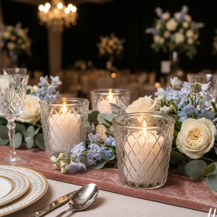 Lit white candles in tipper glass votive holders with blue and white flowers on a pink runner