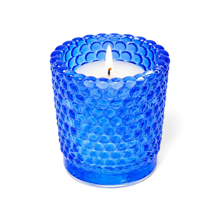 Blue hobnail glass votive candle holder with lit white candle on white background