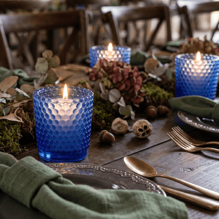 Blue glass hobnail votive holders with glowing candles on rustic floral dining table