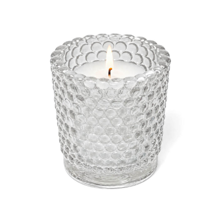A lit white candle inside a clear hobnail glass votive holder on a solid white background