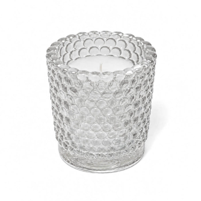 An unlit white candle inside a clear hobnail glass votive holder on a solid white background