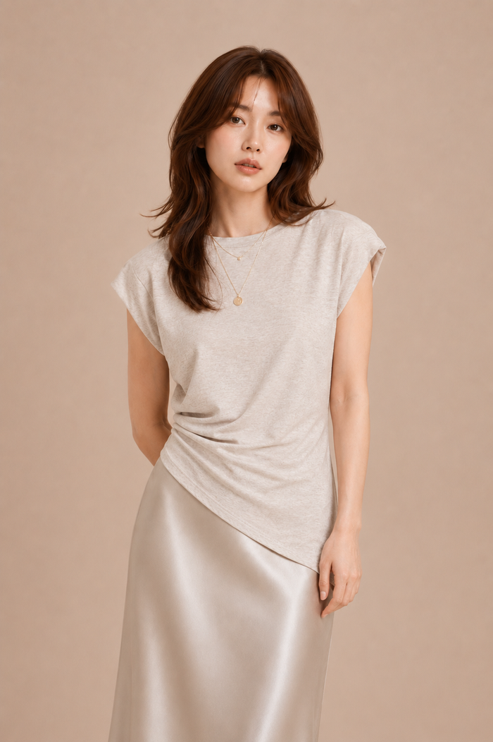 Woman wearing beige asymmetrical top and silk skirt against beige backdrop.