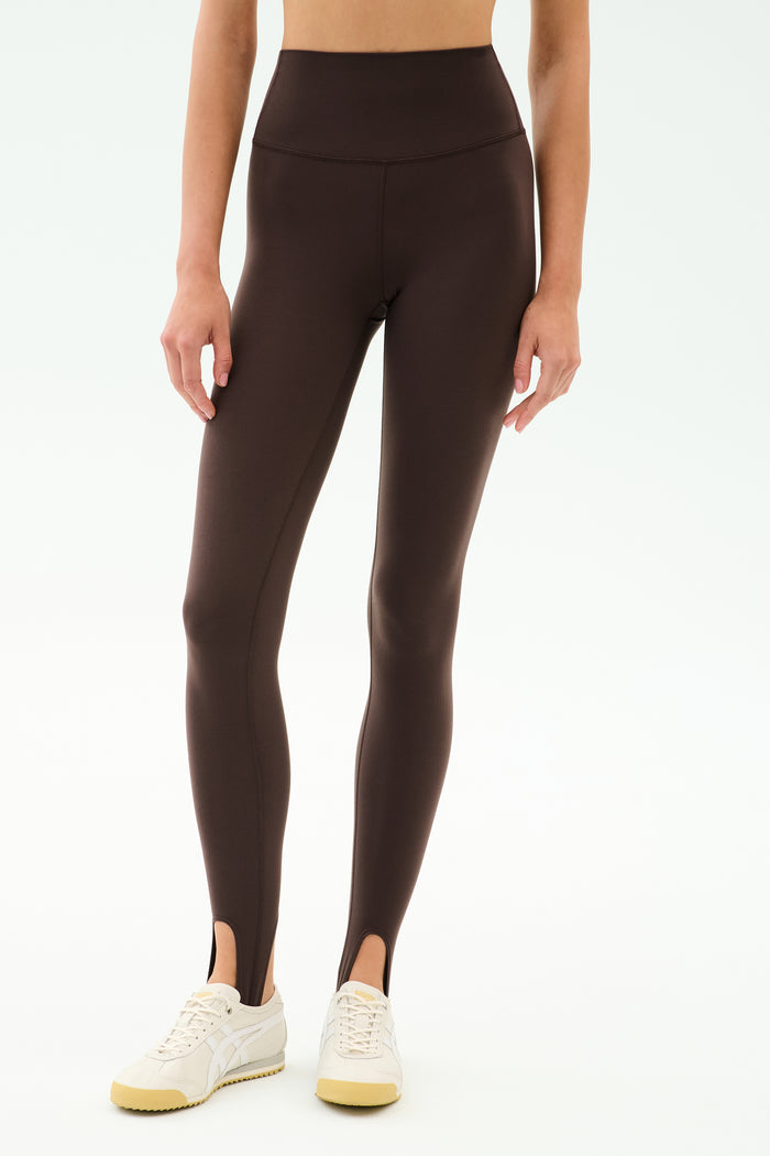 A person wearing SPLITS59 River High Waist Airweight Stirrup leggings in Dark Chocolate, paired with white sneakers, stands against a light background.