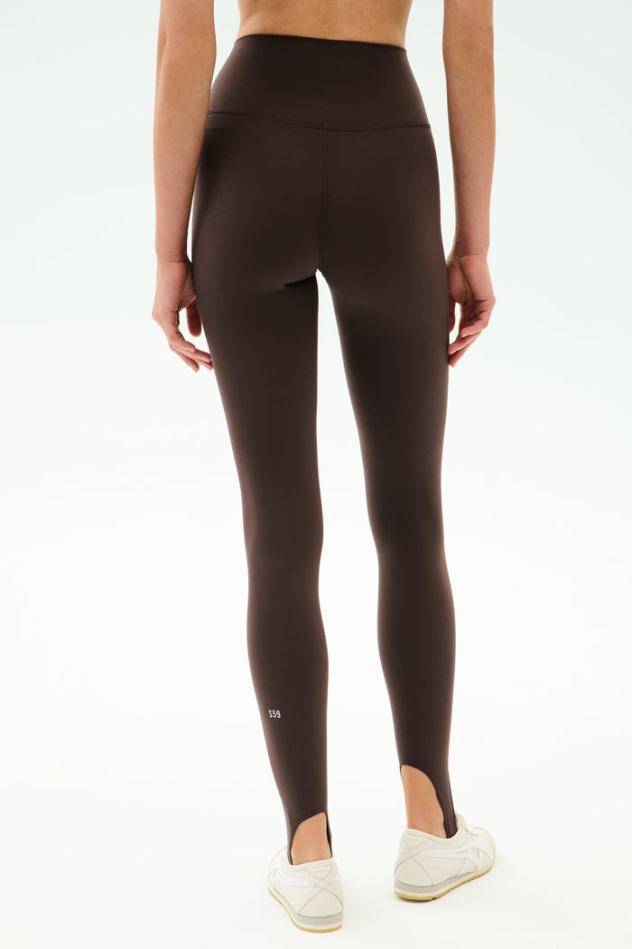 Model wears SPLITS59 River High Waist Airweight Stirrup in Dark Chocolate, styled as stirrup tights with white sneakers, shown from the back against a plain background.