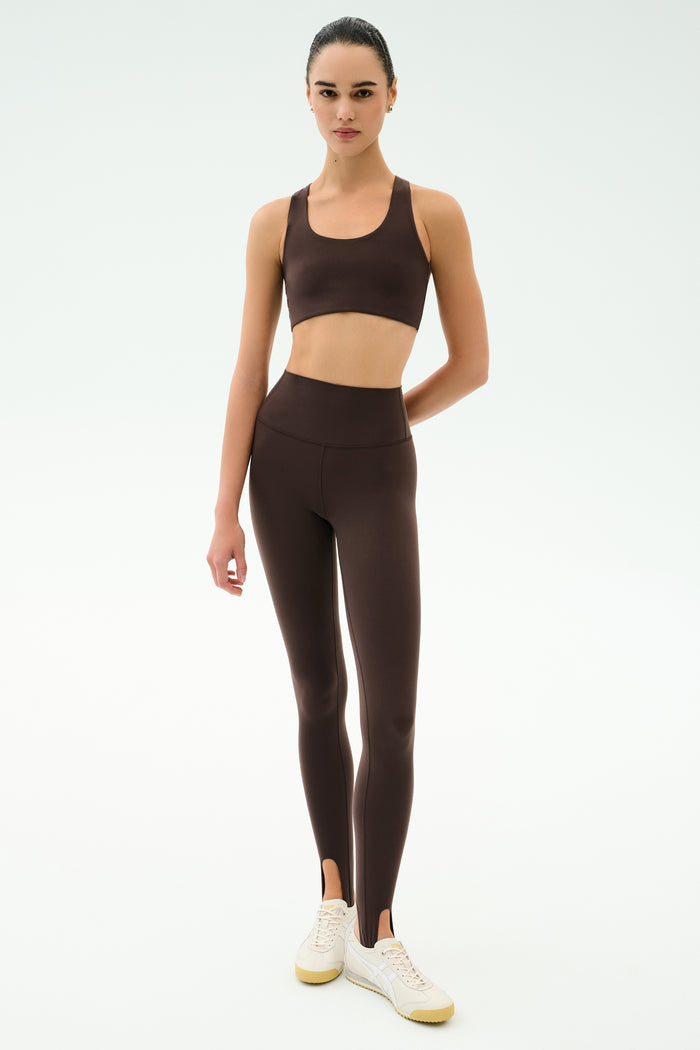 A person stands against a plain background wearing SPLITS59's River High Waist Airweight Stirrup leggings in Dark Chocolate, paired with a matching sports bra and white sneakers.
