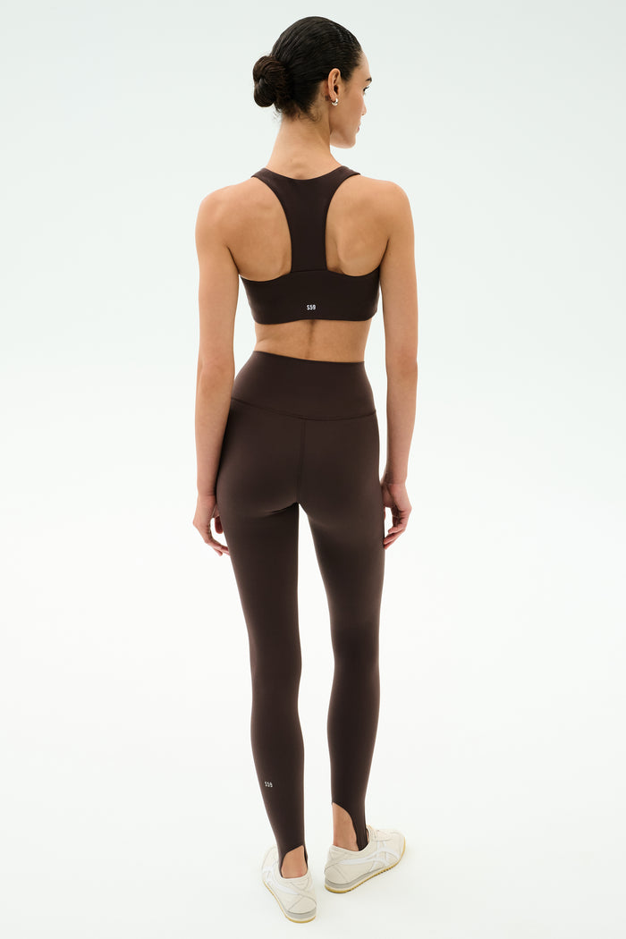 A woman faces away, wearing the SPLITS59 River High Waist Airweight Stirrup leggings in Dark Chocolate with a matching sports bra and white sneakers, set against a plain white background.