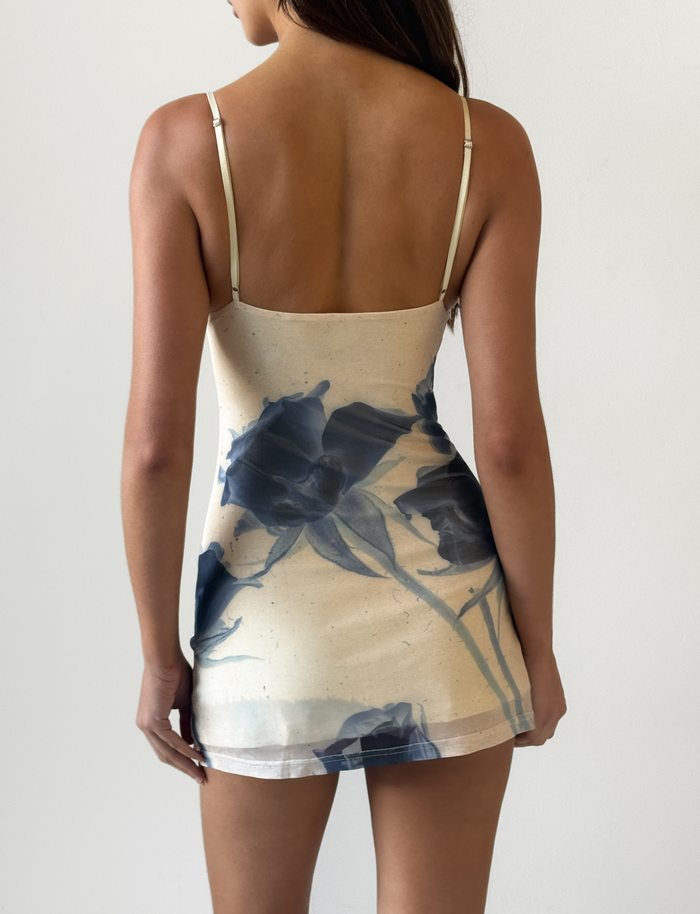 A woman wearing the TIGERMIST ROSY MINI DRESS - INDIGO FLORAL with adjustable cami straps stands facing away from the camera against a plain white background.