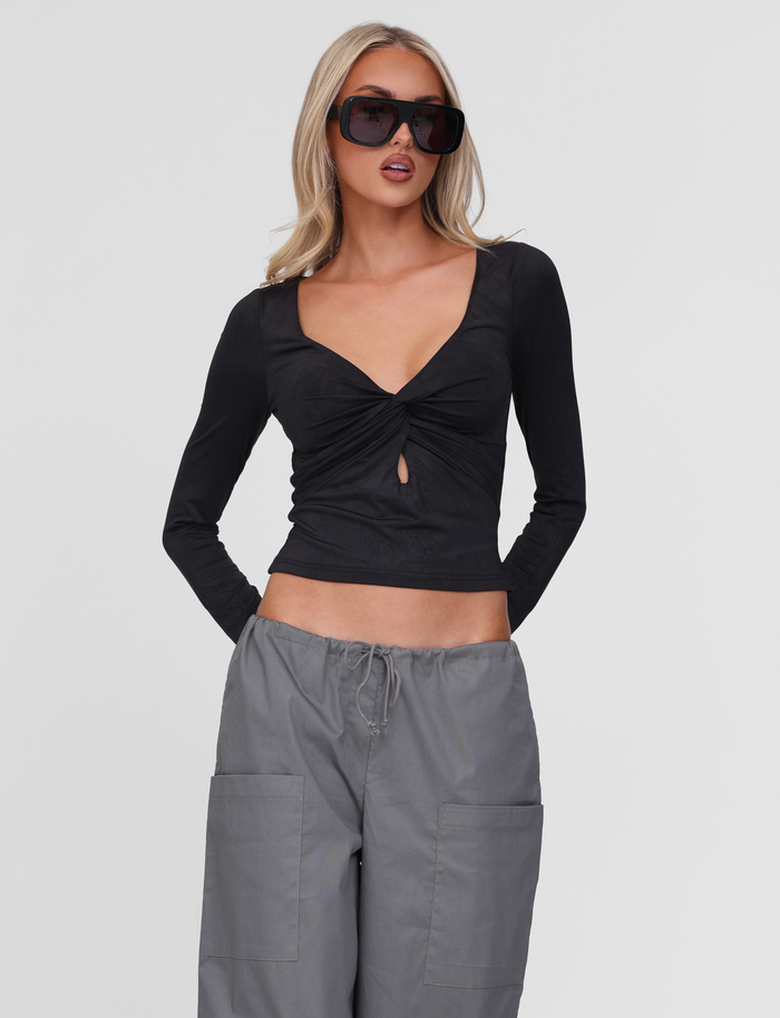 A woman with long blonde hair wears large black sunglasses and the TIGERMIST SAFFIRA LONG SLEEVE TOP in black, featuring a knot and keyhole chest detail, paired with loose gray drawstring pants. She stands against a plain white background.
