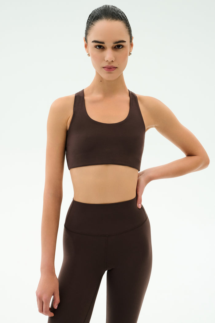 A woman poses against a plain background in SPLITS59’s Sara Airweight Bra - Dark Chocolate and matching leggings, one hand on her hip and a neutral expression—ideal for hot yoga or Pilates.