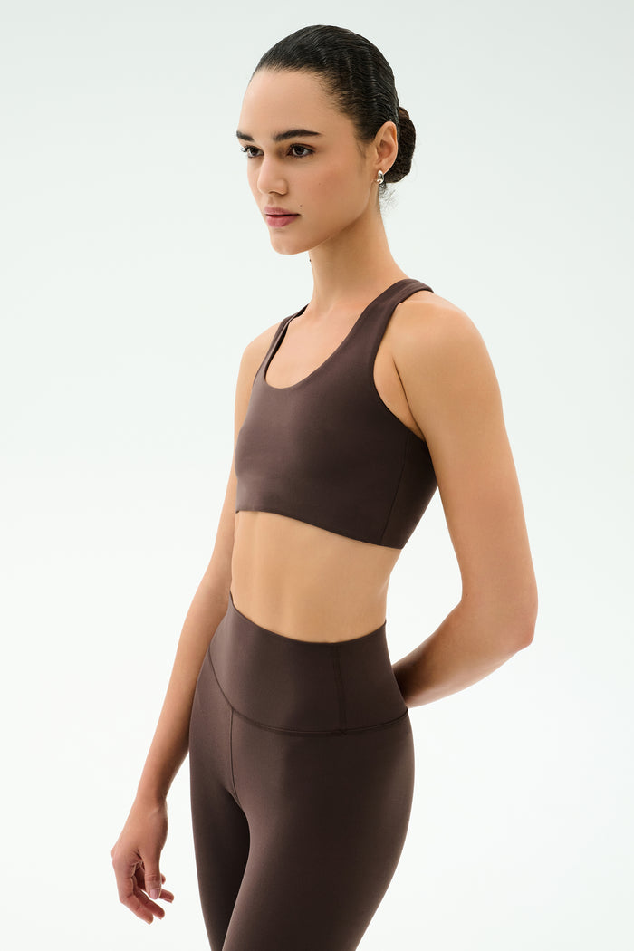 Wearing the SPLITS59 Sara Airweight Bra in Dark Chocolate and high-waisted leggings, someone stands against a plain light background, gazing sideways—an ideal look for Pilates or hot yoga.