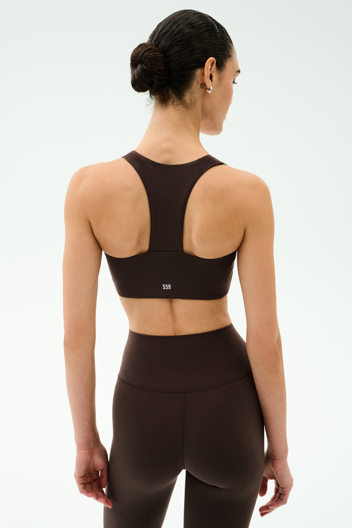 A woman with her hair in a bun faces away, wearing the SPLITS59 Sara Airweight Bra in Dark Chocolate and high-waisted leggings, ready for Pilates against a plain light background.